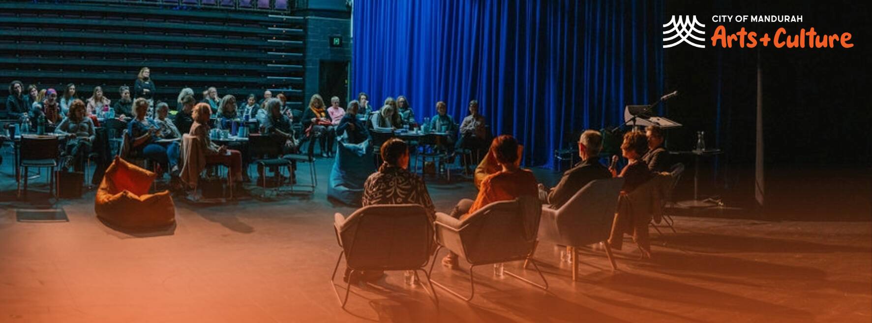 Audience seated in a dark theatre watching a panel discussion on stage.